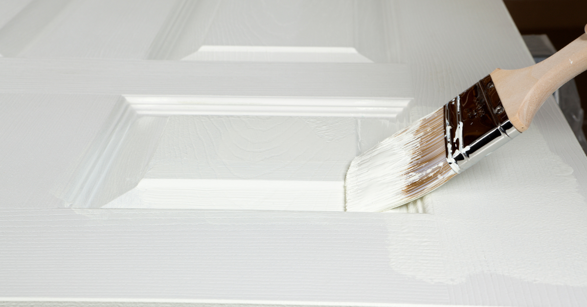 Close-up of a paintbrush applying white bonding primer to a paneled wooden cabinet door for a smooth finish.