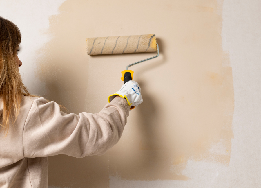 Woman painting an interior wall with a roller while standing beside a step ladder during a residential painting project.