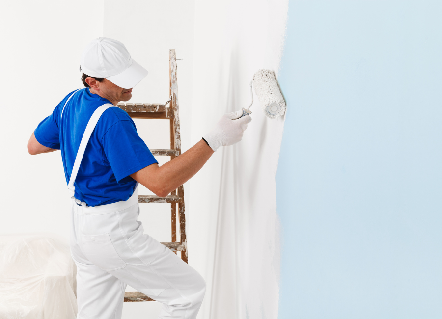 Residential painter in uniform repainting a light blue wall to white using a roller during interior home renovation.
